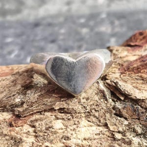 May include: A polished silver-toned ring featuring a prominent heart-shaped centerpiece. The band is smooth and solid, with a reflective metallic surface that catches the light, showcasing a clean, simple design without any additional embellishments.