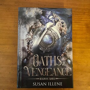 May include: A hardcover book titled Oaths & Vengeance Realm of Zadrya by Susan Illene. The cover features a blue and white bird perched on a silver sword, set against a dark, ornate background with intricate filigree and metallic accents.