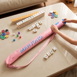 May include: A pink cylindrical carrying case with the word MAHJONG printed in blue, accompanied by a set of white rectangular mahjong tiles with various symbols, several colourful circular game tokens, and small multi-coloured dice.