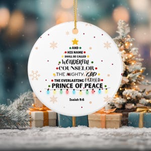 May include: A round white ceramic ornament featuring a gold string, decorated with a Christmas tree graphic formed by the text: AND HIS NAME SHALL BE CALLED WONDERFUL COUNSELOR THE MIGHTY GOD THE EVERLASTING FATHER PRINCE OF PEACE Isaiah 9:6.