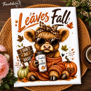 Può includere: Una stampa rettangolare bianca con una mucca highland pelosa con un fiocco leopardato e occhiali da sole, che tiene una tazza di caffè con scritto 'Fueled by CHAOS'. Il testo recita 'Leaves Fall' e 'I DON'T DO BASIC' vicino a zucche e foglie.
