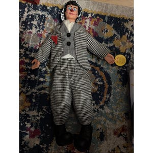 Peut inclure: Une poupée clown vêtue d'un costume pied-de-poule noir et blanc, d'un gilet gris à deux boutons noirs, d'une chemise blanche et d'une pochette rouge. La poupée a un nez rouge, des cheveux foncés, un chapeau rayé noir et blanc et de grandes chaussures noires.