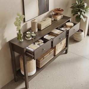 May include: A dark wood console table with a flat top surface, two open drawers, and a bottom shelf. The table holds a glass vase with white flowers, a lantern, a beige radio, books, and candles. Two woven baskets are stored on the bottom shelf.