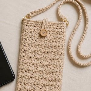 Może przedstawiać: Beżowa szydełkowa torebka na telefon do noszenia przez ramię, wykonana z teksturowanym wzorem ściegu, z zapięciem na jeden drewniany guzik oraz pasującym plecionym paskiem ze sznurka przymocowanym za pomocą złotych metalowych kółek.