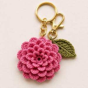May include: A crochet keychain featuring a vibrant pink, multi-layered flower with a textured, petal-like design. Attached to the flower is a small, dark green crocheted leaf. The piece is connected to a gold-toned metal ring and a lobster clasp.