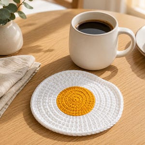 May include: A round, crocheted coaster designed to resemble a fried egg, featuring a bright yellow center surrounded by a textured white border. The handmade textile accessory is circular in shape and displays a consistent, dense stitch pattern.