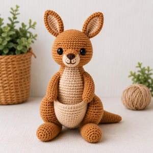 May include: A crocheted kangaroo stuffed animal featuring a medium brown body with a lighter beige belly, chest, and pouch. The toy has large upright ears, black safety eyes, a dark brown stitched nose, and a long tail extending to the right.