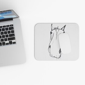 Puede incluir: Una alfombrilla de ratón rectangular blanca con un boceto en tinta negra de una cabeza de caballo de frente. La alfombrilla está situada junto a un portátil plateado con teclado negro y un ratón de ordenador blanco sobre la ilustración.