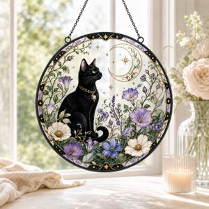 Pode incluir: Um apanhador de sol circular em vitral com um gato preto usando uma coleira dourada com pingente. O gato está rodeado por flores roxas e brancas, vinhas verdes e uma lua crescente com estrelas, tudo dentro de uma moldura de metal preta.
