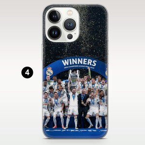 May include: A clear smartphone case featuring a graphic of the Real Madrid soccer team celebrating a victory. The image shows players in white uniforms holding a trophy under confetti, with the text WINNERS UEFA CHAMPIONS LEAGUE FINAL 2024.