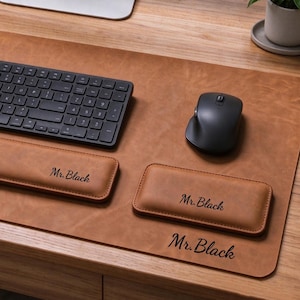 Puede incluir: Un juego de alfombrilla de escritorio de cuero color canela que incluye un reposamuñecas para teclado, uno para ratón y la alfombrilla. Cada pieza tiene grabado el texto Mr. Black en cursiva. Un teclado y un ratón inalámbricos negros están encima.