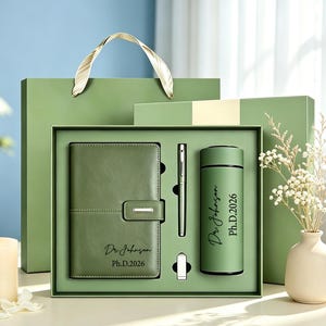 May include: A sage green gift set containing a leather-bound notebook, a metal pen, and a cylindrical vacuum flask. The notebook and flask are personalised with the text Dr. Johnson Ph.D.2026. The items are presented in a matching green box.