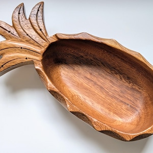 May include: A carved wooden bowl shaped like a pineapple, featuring a natural brown wood grain finish. The bowl has a hollowed-out centre and a detailed top section with carved leaves, showcasing the organic texture and warm tones of the wood.
