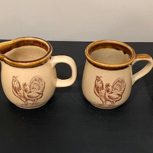 May include: A set of four ceramic kitchen items in a light beige color with brown rims and brown rooster illustrations. The set includes a lidded sugar bowl, a creamer pitcher, and two mugs, all featuring a matching brown chicken design.