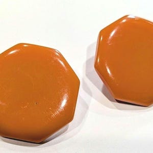 May include: A pair of glossy, burnt orange earrings with a smooth, octagonal shape. Each earring features a flat, solid surface with a reflective finish, showcasing a uniform color throughout the design. The earrings are presented as a matching set.