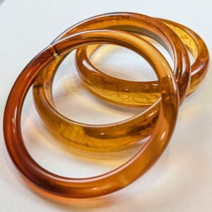 May include: Three circular bangles crafted from a translucent, amber-colored material. Each bangle features a smooth, polished surface with swirling, marbled patterns in varying shades of golden brown and deep orange, creating a layered effect.