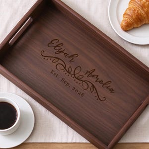May include: A rectangular dark brown wooden serving tray with cutout handles. The center features laser-engraved text reading "Elijah & Amelia" with a decorative flourish and the date "Est. Sep, 2026" below the names.