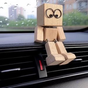 Può includere: Una piccola figura di robot in legno con testa e corpo a forma di cubo, arti rettangolari e occhi scuri in filo metallico a forma di infinito. La figura è costruita con blocchi di legno chiaro collegati da giunti in filo metallico flessibile.