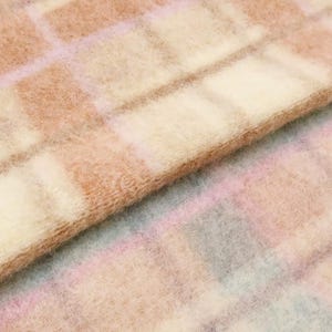 Op de afbeelding: Een gevouwen stuk stof met een zachte, pluizige textuur en een grootschalig ruitpatroon. Het ontwerp bestaat uit elkaar kruisende lijnen in tinten lichtroze, crème en beige, die een raster van vierkanten op het oppervlak vormen.
