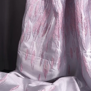 May include: A fabric with a white base and a raised, textured pattern of light pink, flame-like or leaf-shaped motifs. The material has a subtle sheen and a structured, slightly stiff appearance, with the pink elements creating a repeating design.