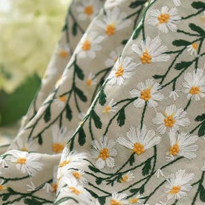 May include: A light beige fabric featuring an embroidered pattern of white daisies with yellow centers and dark green stems and leaves. The fabric is draped in soft folds, showcasing the detailed floral stitching across the surface.