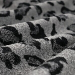 May include: A soft, textured fabric featuring a repeating leopard print pattern in shades of charcoal black and heather gray. The material is draped in gentle folds, highlighting the fuzzy, tactile surface of the animal-inspired textile design.