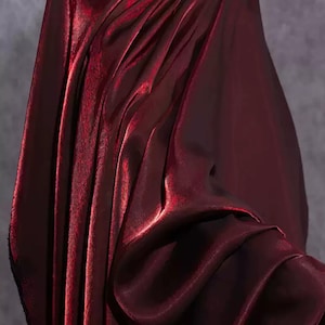 Pode incluir: Um tecido bordô profundo com um brilho metálico e uma superfície lisa e reflexiva. O material está drapeado em dobras verticais, criando luzes e sombras que enfatizam a textura e a rica cor vermelha escura do material têxtil.