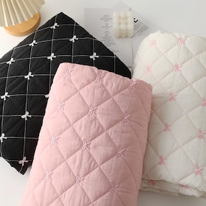 May include: Three quilted fabric pieces in black, light pink, and white. Each piece features a diamond-stitched pattern with small embroidered bows at the intersections. The black fabric has white bows, the pink has pink bows, and the white has pink bows.