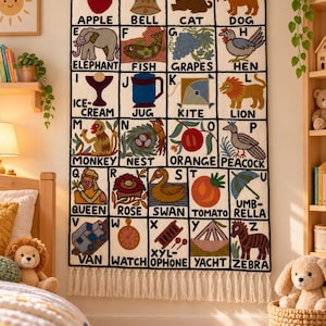 May include: A rectangular wall hanging featuring a grid of the alphabet from A to Z, each letter paired with a corresponding illustration and word. The textile piece has a cream background, black grid lines, and a white fringe along the bottom edge.