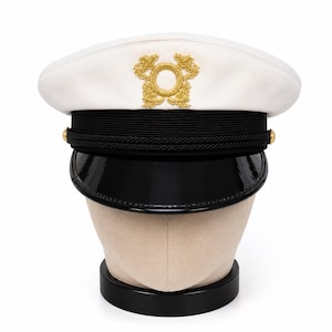 May include: A white peaked cap featuring a black band and a glossy black visor. The front of the white crown displays an ornate gold embroidered emblem. A thin black cord runs across the front of the band, secured by small gold-toned buttons.