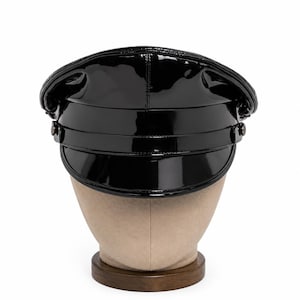 Puede incluir: Una gorra de estilo militar hecha de cuero de charol negro con un acabado de alto brillo. El sombrero presenta una corona estructurada, una visera ancha y una banda horizontal en la parte delantera asegurada por dos botones circulares a cada lado.