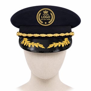 Op de afbeelding: Een marineblauwe kapiteinspet met een goudkleurig gevlochten koord aan de voorzijde en een zwarte klep met gouden bladerborduurwerk. Een ronde badge op de voorkant toont een kroonicoon boven de tekst YOUR LOGO OR TEXT HERE en een kleine ster.