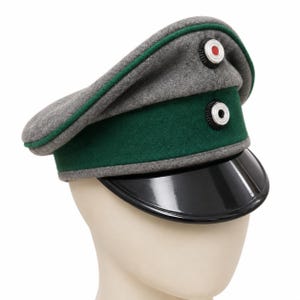 Puede incluir: Una gorra militar de lana gris con una banda verde oscuro, una visera negra brillante y dos escarapelas circulares. La escarapela superior tiene un centro rojo, mientras que la inferior tiene un centro blanco, ambas rodeadas por anillos negros y plateados.