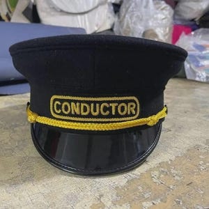 May include: A black conductor cap featuring a flat top, a stiff black visor, and a gold embroidered patch on the front with the word CONDUCTOR in capital letters. A decorative gold braided cord is attached across the front of the cap.