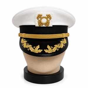 Op de afbeelding: Een wit-zwarte marineofficierspet met een gouden gevlochten kinband en goudkleurig eikenbladborduursel op de zwarte klep. De voorzijde van de witte kroon toont een vierkant embleem met een cirkelvormig goudkleurig geborduurd symbool.