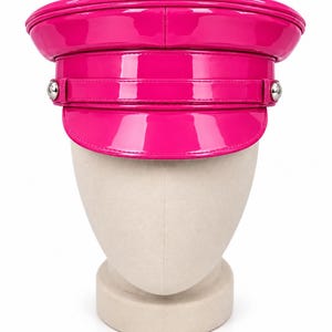 May include: A bright hot pink patent leather military-style cap with a glossy finish. The hat features a structured flat top, a curved visor, and a horizontal band across the front secured by two small, round, silver-toned metal buttons on each side.