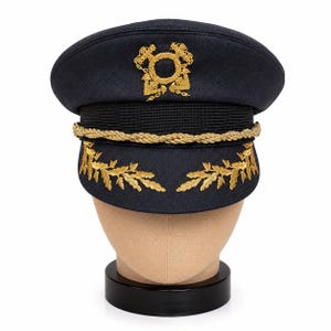 Op de afbeelding: Een donkerblauwe pet met klep en gouden borduursels aan de voorzijde. De pet is voorzien van een goudkleurig gevlochten koord over de band, een centraal goud geborduurd embleem en gouden bladmotieven op de klep, geplaatst op een neutrale basis.