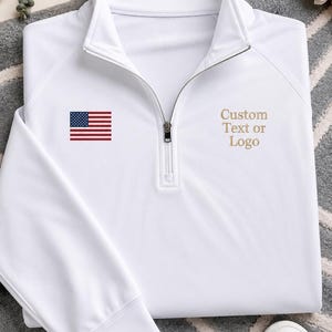 Peut inclure: Un pull blanc à manches longues et fermeture éclair au quart, doté d'un petit drapeau américain brodé sur la poitrine droite et du texte "Custom Text or Logo" brodé en couleur beige sur la poitrine gauche, avec une tirette de fermeture éclair en métal argenté.