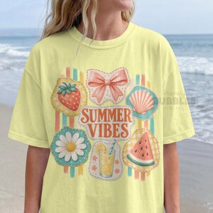 Puede incluir: Una camiseta de manga corta de color amarillo pálido con un gráfico central que incluye el texto "SUMMER VIBES" en letras naranjas. El diseño presenta ilustraciones de una fresa, un lazo rosa, una concha, una margarita, una limonada y una sandía.