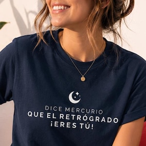 Puede incluir: Una camiseta azul marino de manga corta con un gráfico blanco de una luna creciente y estrellas sobre el texto "DICE MERCURIO QUE EL RETRÓGRADO ¡ERES TÚ!". La camiseta tiene cuello redondo y se combina con pantalones vaqueros azul claro y zapatillas.