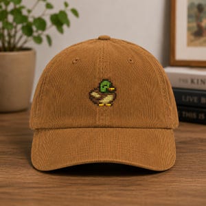 May include: A brown corduroy baseball cap featuring a small, centered pixel art embroidery of a mallard duck. The duck design displays a green head, yellow beak, and brown body, rendered in a blocky, digital-inspired style on the front panel.