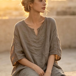 May include: A loose-fitting, taupe-colored tunic with a V-neckline and three-quarter length sleeves. The garment features a raw, frayed hemline along the edges of the sleeves and the bottom, crafted from a textured, lightweight fabric.