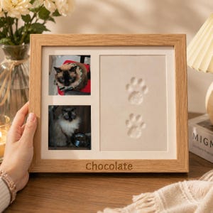 May include: A light wood rectangular picture frame featuring two square photo slots on the left side containing images of a Siamese cat and a larger rectangular space on the right with two embossed paw prints. The name Chocolate is engraved.