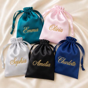 May include: Five satin drawstring pouches in teal, light pink, white, black, and navy blue. Each pouch features gold embroidered names: Emma, Olivia, Sophia, Amelia, and Charlotte. The bags have matching satin ribbon drawstrings at the top.