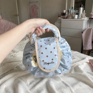May include: A light blue drawstring pouch with a brown polka dot pattern and a beige trim. The front flap features the word Magnolia embroidered in dark brown script. The bag has a rounded shape with a top handle and a gathered yellow interior lining.