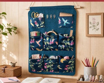 May include: A dark blue wall pocket organizer with a floral and hummingbird print, featuring eight pockets hanging from a wooden dowel. The text reads Sami Dolls, PDF Sewing Pattern, and WALL POCKET ORGANIZER, with icons for download and printing.