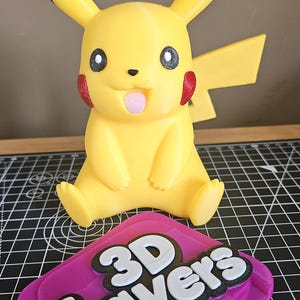 May include: A yellow Pikachu figurine with black-tipped ears, red cheeks, and a pink tongue, positioned behind a purple, black, and white 3D-printed plaque featuring the text 3D Layers in a bold, layered font style on a black grid mat.