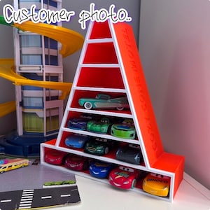 May include: A bright orange, cone-shaped display shelf with white trim and multiple horizontal tiers. The side of the structure features the embossed text COZY CONE MOTEL. The shelves are filled with various small, colourful toy race cars.