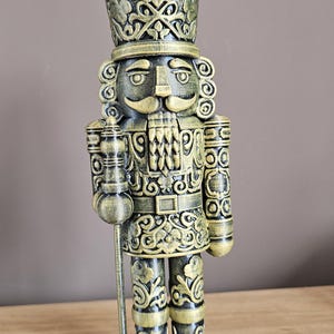 May include: A decorative nutcracker figurine featuring a metallic gold and black finish with intricate embossed scrollwork patterns. The figure wears a tall hat, has a prominent moustache, and holds a long staff in its right hand.