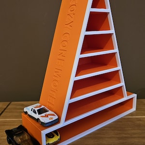 May include: An orange and white display shelf shaped like a traffic cone with the text COZY CONE MOTEL embossed on the side. The shelf features multiple horizontal tiers for storage. Two small toy cars are positioned on the bottom level and base.